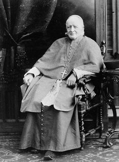 Black and white portrait of Bishop Ignace Bourget seated on a chair in a living room. He is wearing religious attire: a cassock, white skullcap and a cross around his neck.