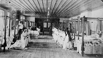 Black and white photo of a hospital room. There are two series of curtained beds along the walls with patients in each. Nuns kneel at the foot of the beds, praying and listening to the prayer of a priest in the room.