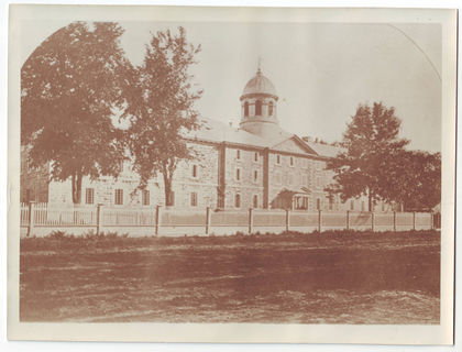 Large rectangular stone building with many windows. The building is partially hidden by trees and a fence.