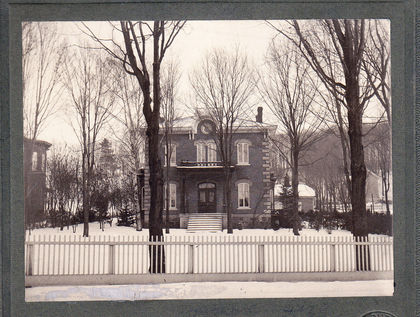 Photo of the front of the Laurier residence. A two-storey brick home with large windows and a balcony overlooking the entrance. The property is bordered by a fence and small bushes.