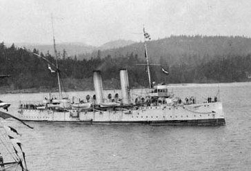 Black and white photo of a metal ship with a mast at each end and two chimneys at the centre, towing a small wooden boat behind. Behind the ship is the great forest that characterized the port of Prince Rupert in 1914.