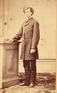 Sepia photo of Wilfrid Laurier, a student at Collège de l'Assomption. He stands to the side with his right arm on a pedestal and is looking at the camera. He wears a long jacket down to the thighs, a shirt with a white collar, a tie. black trousers and black shoes.