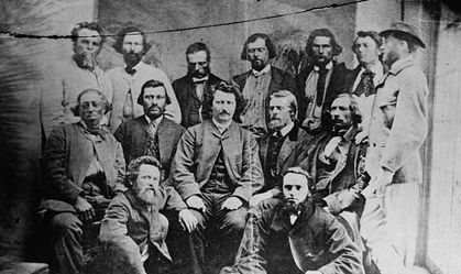 Photographie d'un groupe d'homme Métis du Manitoba, faisant partie du gouvernement de la Nation Métis. Assis au centre du groupe se trouve Louis Riel les mains sur les cuisses, portant un habit et regardant le photographe.