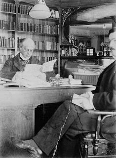 Black and white photo of Laurier seated at his desk reading a document. Behind him, a large bookcase and a mirror. Facing him, a man seated in a chair (Mr. Boudrias).