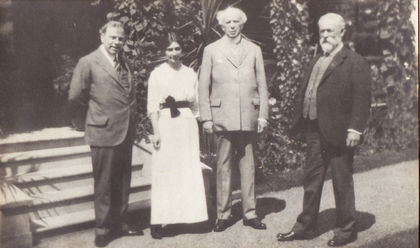 Black and white photo taken at the home (in the background) of the Honorable Sydney Fisher. From right to left: the Honorable Sydney Fisher in a black suit; W. Laurier in a grey suit, holding a cane; Mrs. Arthur Branchaud in a white dress and Mackenzie King in a grey suit.