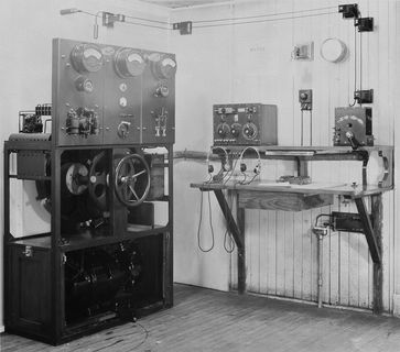 The installation consists of radio equipment to the right with two headsets and a radio receiver. To the left is a large piece of equipment with a wheel at its centre, dials and a lever above and mechanical equipment below. Cables hung from the top of the wall connect the two components.