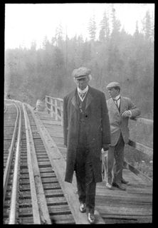 Black and white photo of a railway alongside a forest. Wilfrid Laurier is in the foreground wearing a coat and a hat. He is walking on a wooden walkway along the railway. Behind him is a man wearing a grey suit, looking to his left.