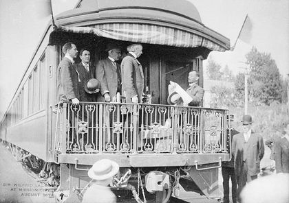 Black and white photo of Laurier and a group of men standing on the rear balcony of a railway car. Another man standing on the railway car's stairs is facing Laurier and reading a text. A crowd surrounds the car.
