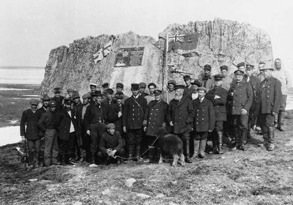 Black and white photo of Captain Joseph-Elzéar Bernier, his crew and a sheep assembled for a group photo. Behind him is a huge rock on which is installed a commemorative plaque and the Canadian flag at the time. The ocean lies behind the huge rock.