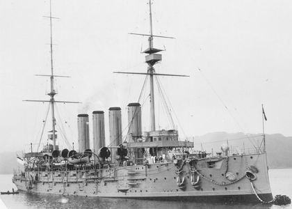 Black and white photo of a two-masted metal ship with four chimneys. The ship is docked and there is a small wooden boat near it carrying a man who is talking to another man on the Niobe. On the side of the ship are its anchors and lifeboats. There is also a cabin of sorts built around the masts.