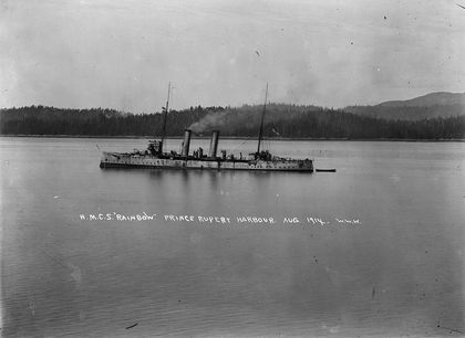 Black and white photo of a metal ship with a mast at each end and two chimneys at the centre towing a small wooden boat behind. Behind the ship is the forest forest that characterized the port of Prince Rupert in 1914.