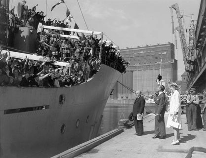 A man and a woman on a wharf greeting a large group of men and women on a metal ship.