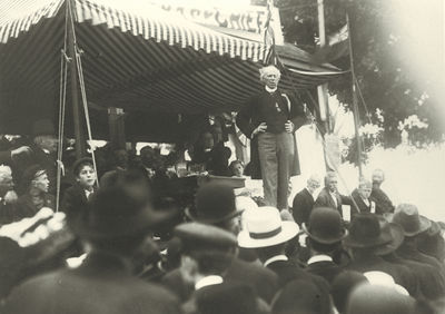 White-haired man talking on a promontory in front of a crowd