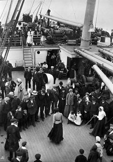 Group of men, women and children on a ship's deck. Some form a circle to watch a woman skip rope. They are passing the time during the trip to Canada.