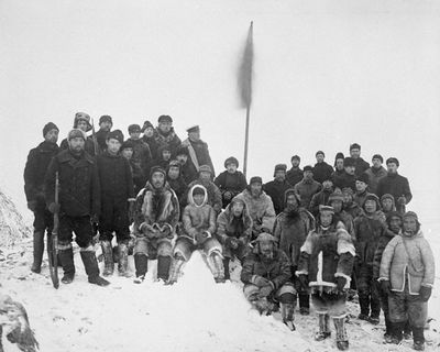 Photographie en noir et blanc, représentant le capitaine Joseph-Elzéar Bernier et son équipage, devant un drapeau hisser sur un poteau. Il se trouve sur un monticule de roche et de neige.