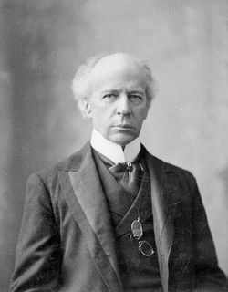 Older man with white hair, wearing a black suit with a white collar, looking directly at the camera lens.